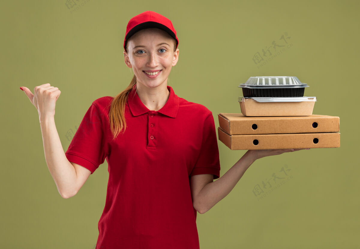 自信身穿红色制服 头戴鸭舌帽的年轻送货女孩 手里拿着披萨盒和食品包 自信地微笑着指着绿墙那边壁板盒子年轻人