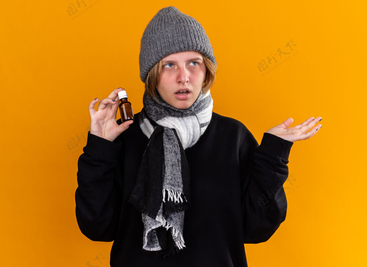 周围不健康的年轻女子戴着暖和的帽子 脖子上围着围巾 感觉不舒服 患了感冒和流感 手里拿着药瓶 胳膊站在橙色的墙上介绍瓶子生病