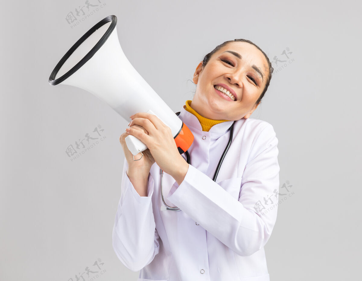 医生身着白色医用外套 脖子上戴着听诊器 手持扩音器的年轻女医生高兴地站在白墙上 脸上洋溢着乐观的微笑听诊器脖子外套