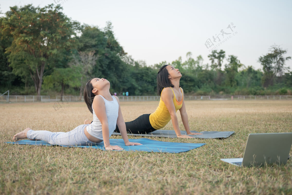 锻炼妈妈和女儿在做瑜伽在公园里训练妇女和孩子户外运动健康的运动生活方式 看瑜伽练习在线视频教程和伸展胸部和脊柱伸展视频体育