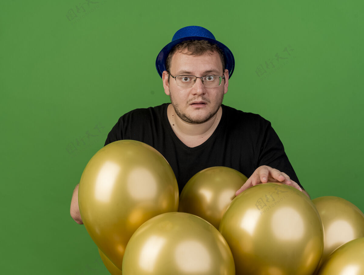 光学焦急的成年斯拉夫人戴着眼镜 戴着蓝色的派对帽 站着氦气球绿色穿成人