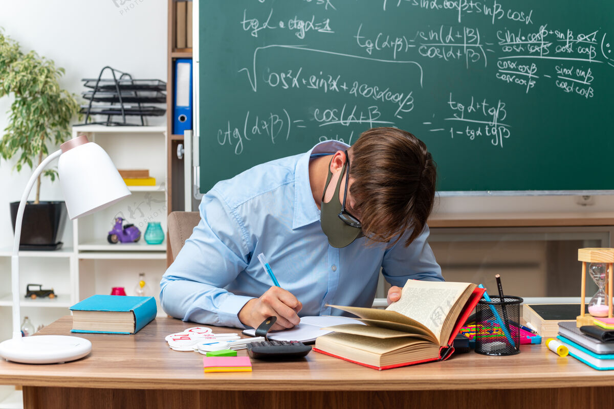 坐着年轻的男老师戴着眼镜和保护面具写备课坐在课桌前在教室里用黑板前的书和笔记准备数学穿