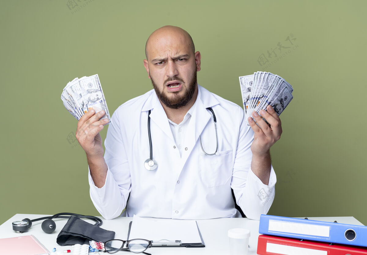 年轻愤怒的年轻秃头男医生穿着医用长袍和听诊器坐在办公桌旁听诊器工具工作