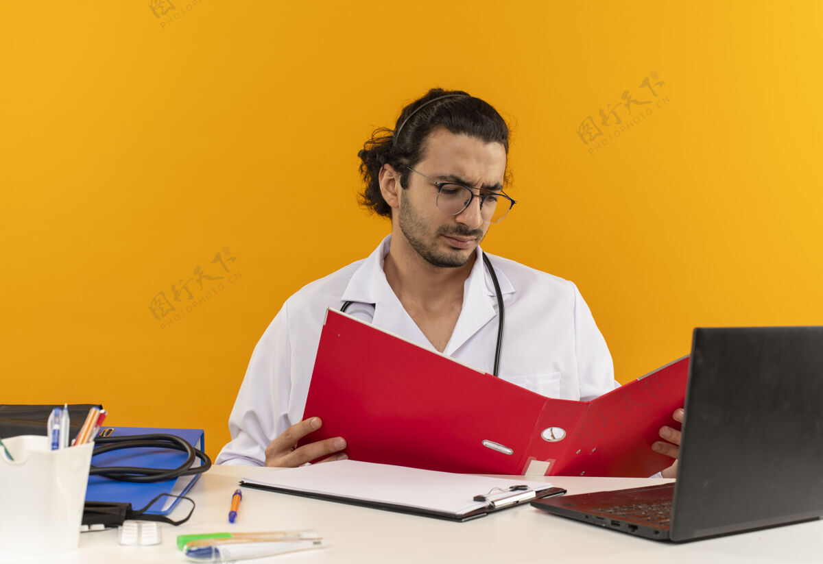 复制年轻的男医生戴着医用眼镜 穿着医用长袍 听诊器坐在办公桌旁听诊器文件夹空间