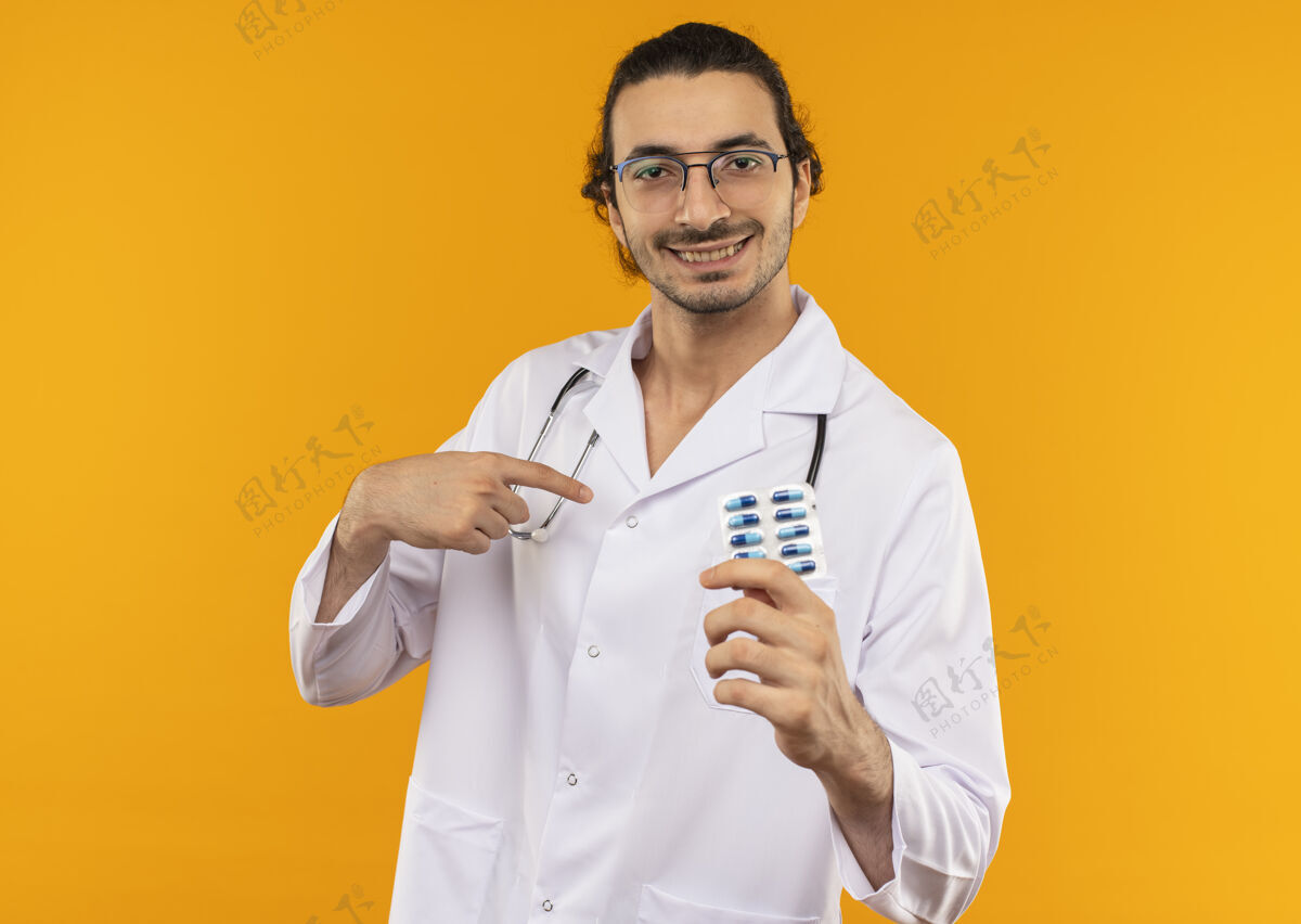黄色微笑的年轻医生戴着医用眼镜穿着医用长袍手持听诊器拿着药片指着自己微笑空间持有