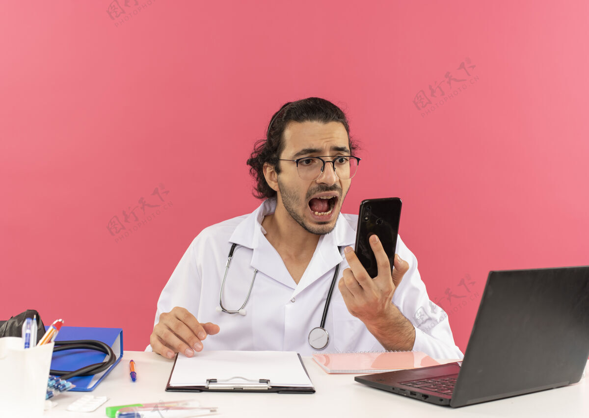 害怕戴着医用眼镜 穿着医用长袍 带听诊器的年轻男医生坐在办公桌旁 吓了一跳穿工作工具