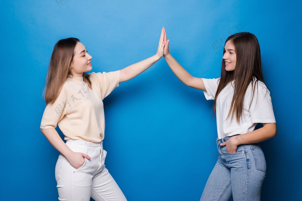 指着两个长着不同头发的快乐的年轻女子隔着蓝色的墙互相击掌笑欢呼女人