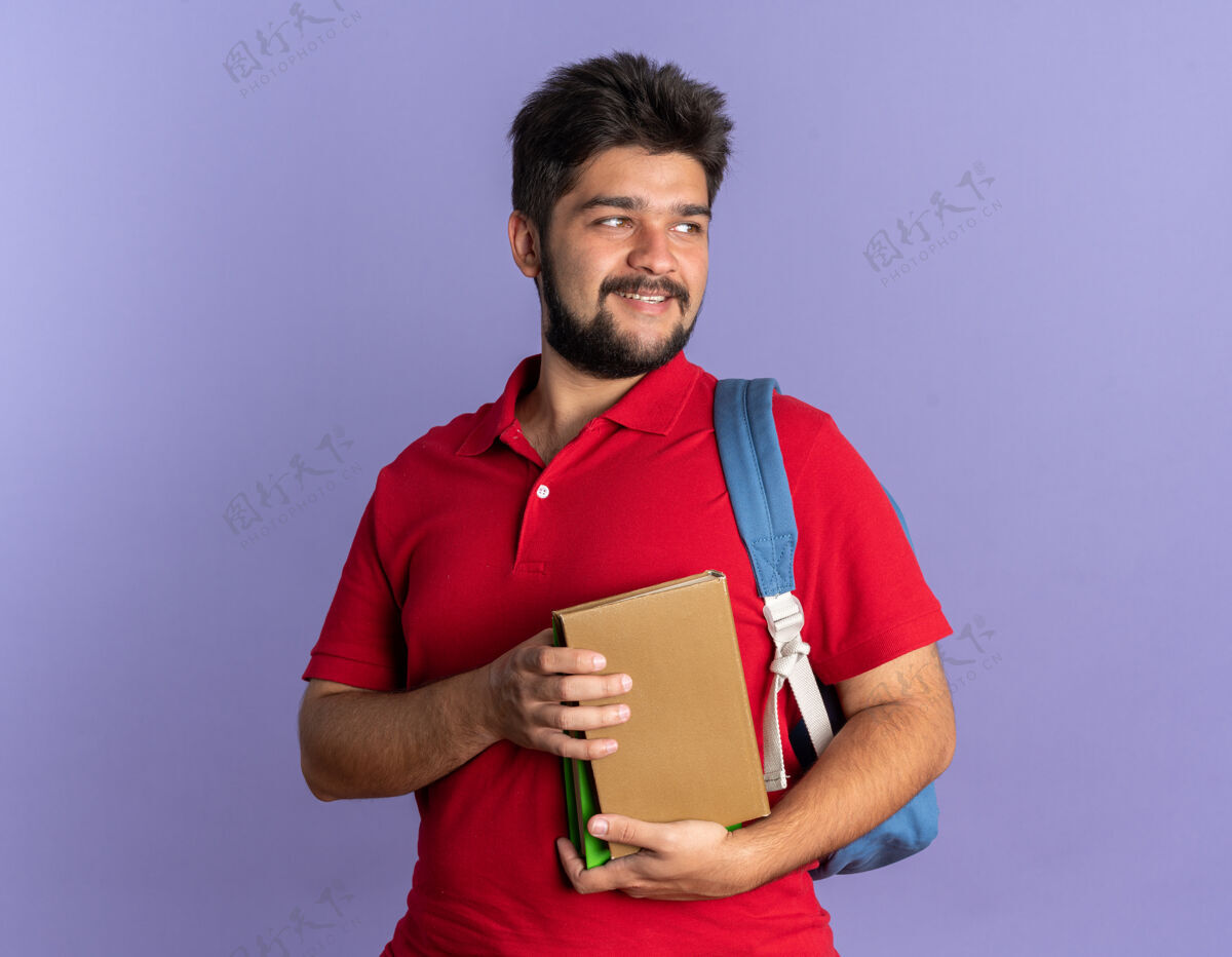 微笑年轻的留着胡子的学生 穿着红色马球衫 背着书包 面带微笑 站在一边看着书抱着快乐学生