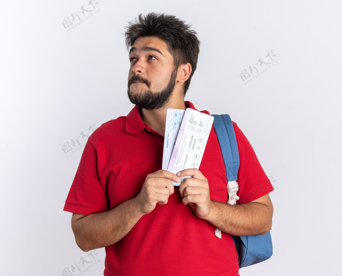小伙子年轻的留着胡子的学生 穿着红色马球衫 背着背包 手里拿着机票 面带微笑 抬头看去 站在那里积极思考马球微笑正面