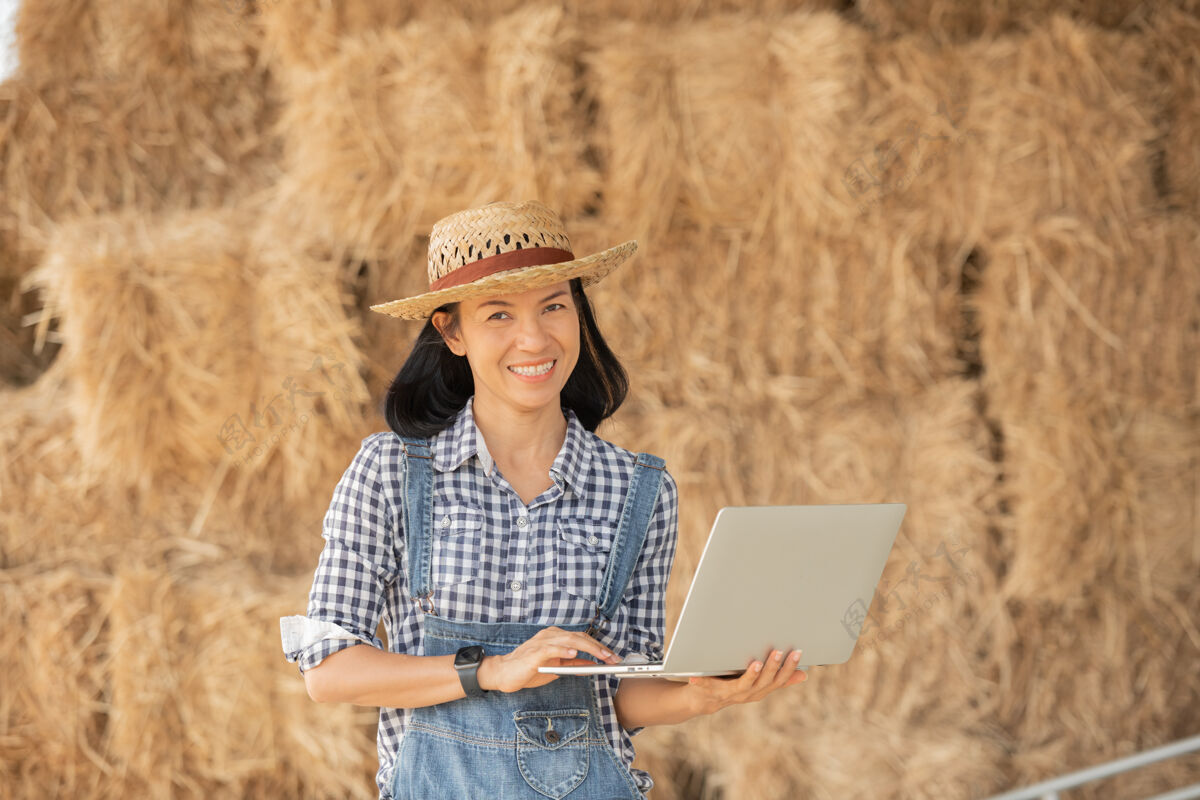 数据亚洲年轻的女农民戴着帽子站在地里 在笔记本电脑键盘上打字带着笔记本电脑的妇女监督农田 概念生态 交通 清洁空气 食品 生物制品的工作自然电脑女人