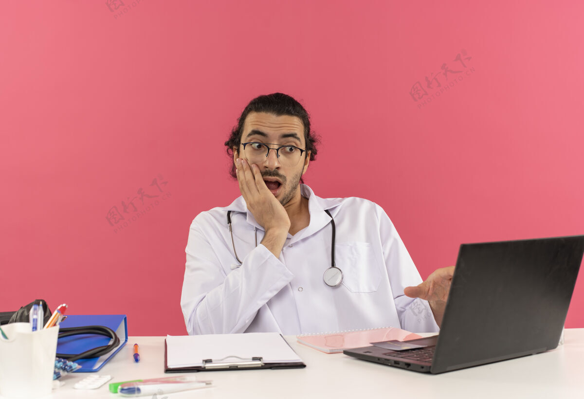 空间惊讶的年轻男医生戴着医用眼镜 穿着医用长袍 听诊器坐在办公桌旁长袍工具医疗