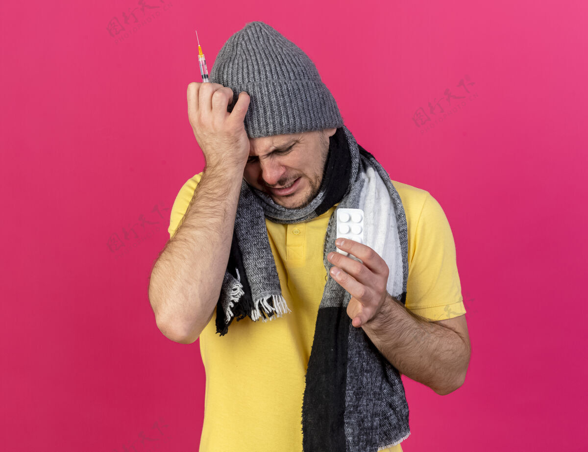 药片疼痛的年轻金发病斯拉夫男子戴冬季帽子和围巾把手放在头上举行注射器帽子斯拉夫医疗