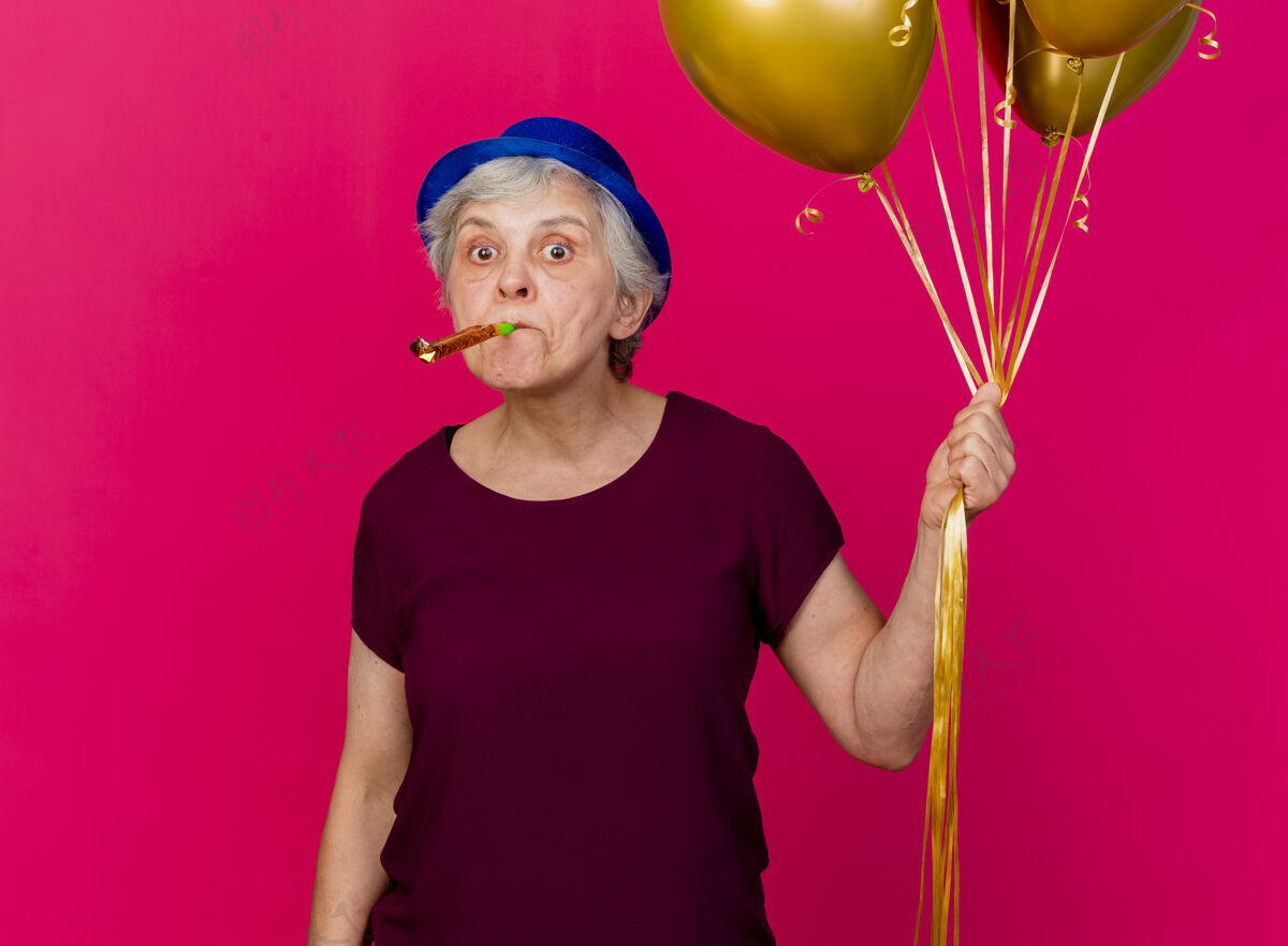 穿惊讶的老妇人戴着聚会帽手持氦气球吹着粉红色的口哨气球惊喜粉色