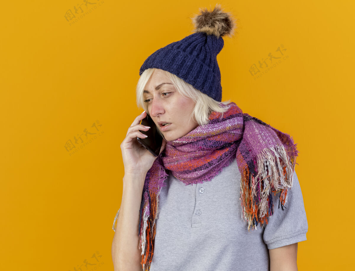 疾病苍白的年轻金发病斯拉夫妇女戴着冬天的帽子和围巾在电话上交谈孤立的橙色围巾女人金发