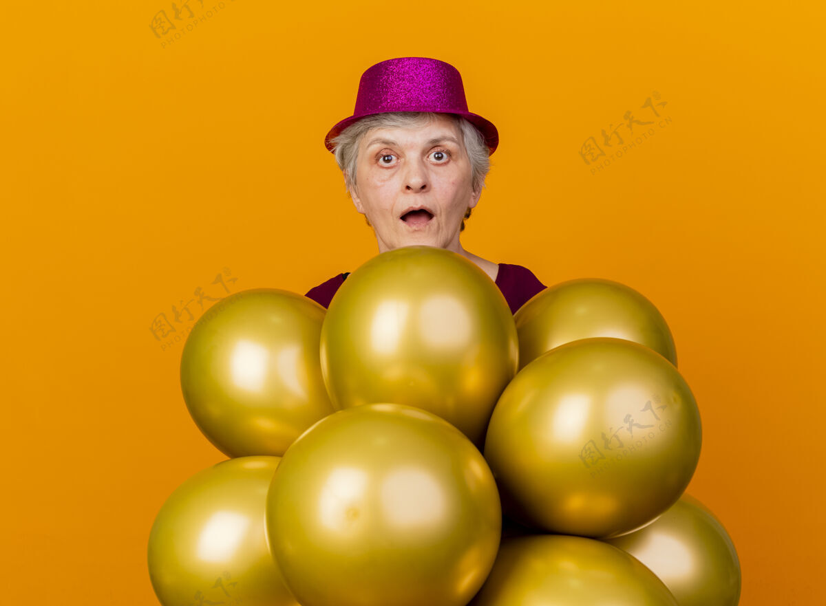惊喜惊讶的老妇人戴着派对帽站在橙色的氦气球上氦帽子女人