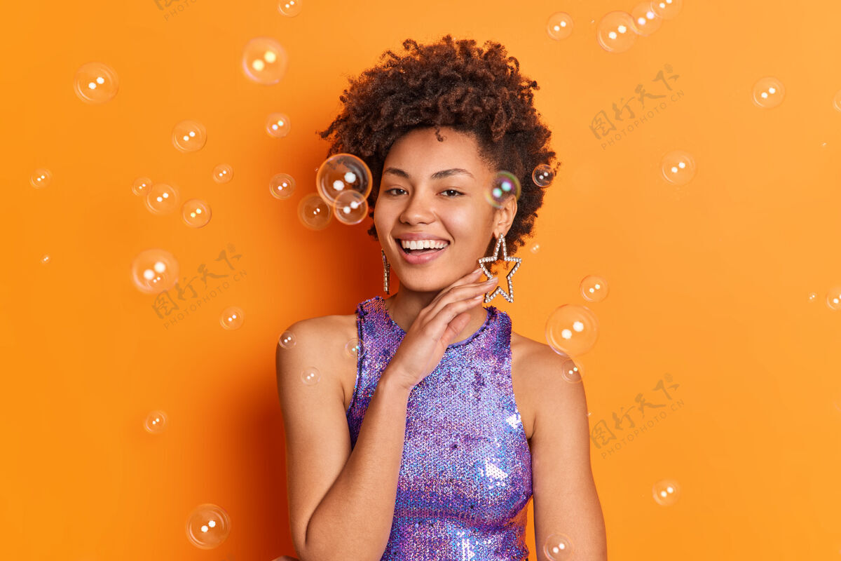 头发时尚开朗的非裔美国女士轻轻触碰下巴线条有时髦的发型微笑宽大地穿着时髦的闪闪发光的紫色衬衫星形耳环摆姿势在橙色的墙上肥皂泡周围美丽时尚脸