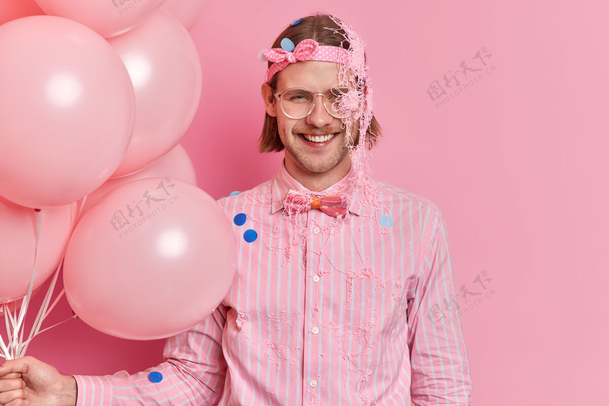 涂抹快乐的欧洲成年男子戴着头带和条纹衬衫 领结上沾满了蛇纹石喷雾 享受着派对庆典 手里拿着一堆气球 孤立在粉红色的墙上请事件举行