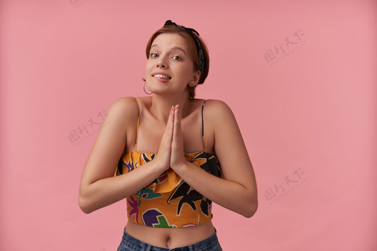 成人可爱俏皮的年轻女子 穿着五颜六色的上衣 戴着头巾 双手抱在祈祷的姿势 在粉色的墙上咬着嘴唇宗教可爱拍摄