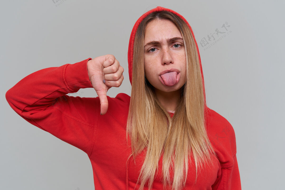 时尚悲伤沮丧的金发女郎 穿着红色连帽衫 在灰色的墙上露出舌头和大拇指站立模特情感
