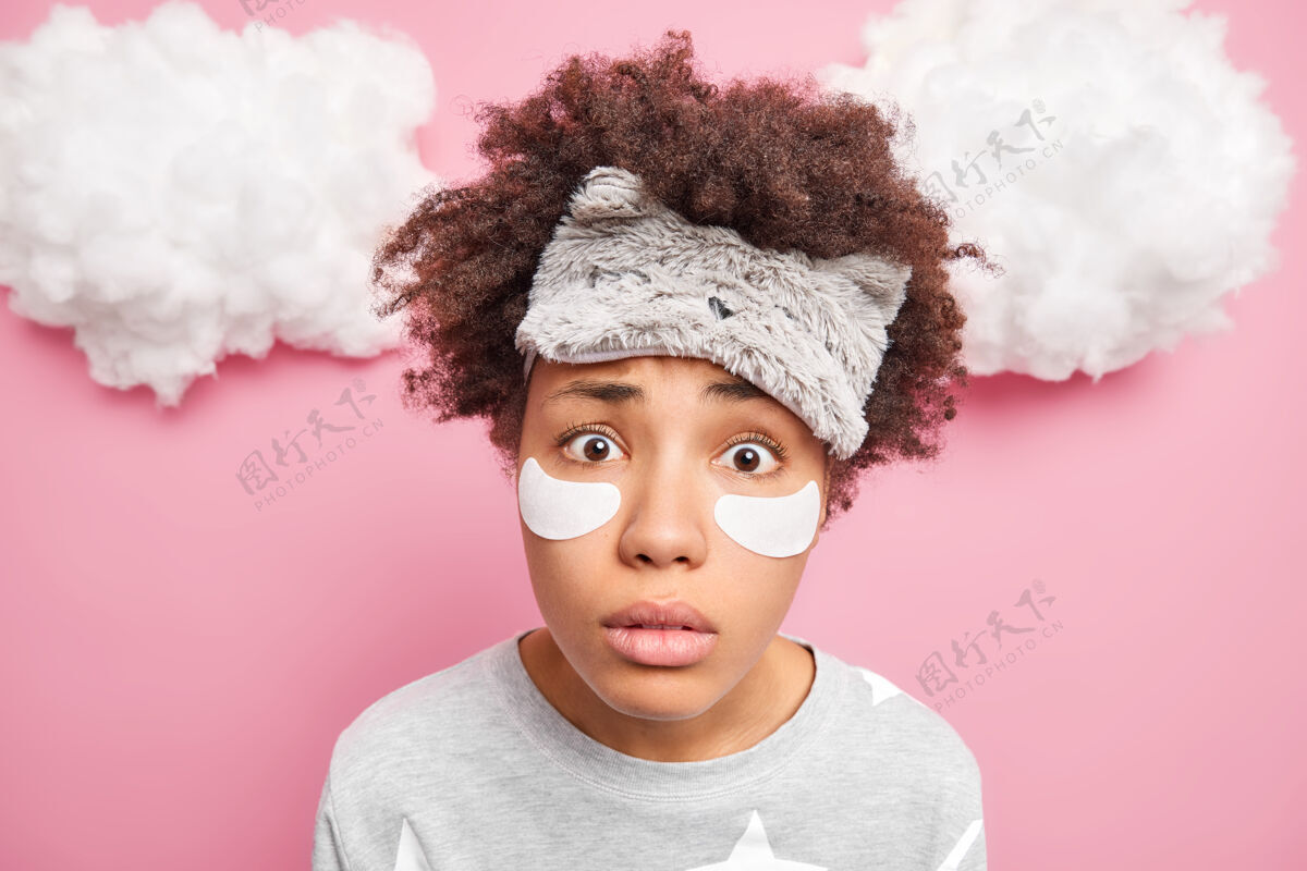 头发特写镜头中 一位女士惊讶地盯着相机 戴着睡眠面罩 穿着隔离在粉色墙壁上的睡衣 在眼睛下面贴上眼罩 以减少皱纹醒来贴片放松