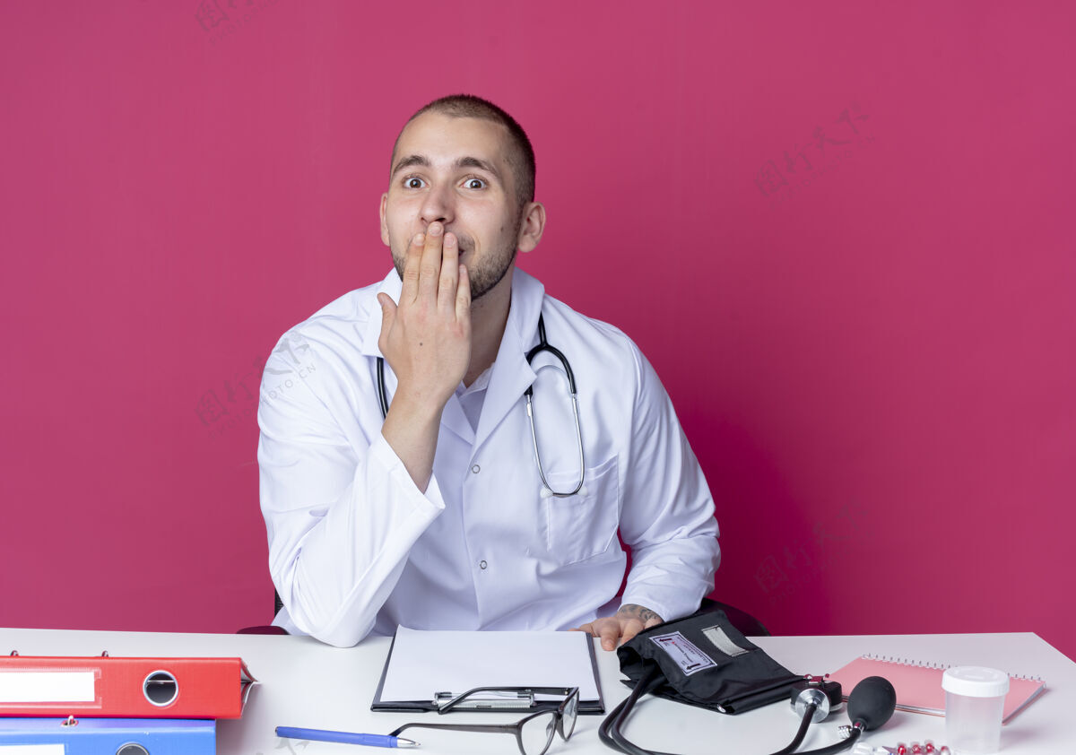 感觉惊讶的年轻男医生穿着医用长袍和听诊器坐在办公桌旁 手放在隔离在粉红色墙上的嘴巴上姿势脸年轻人