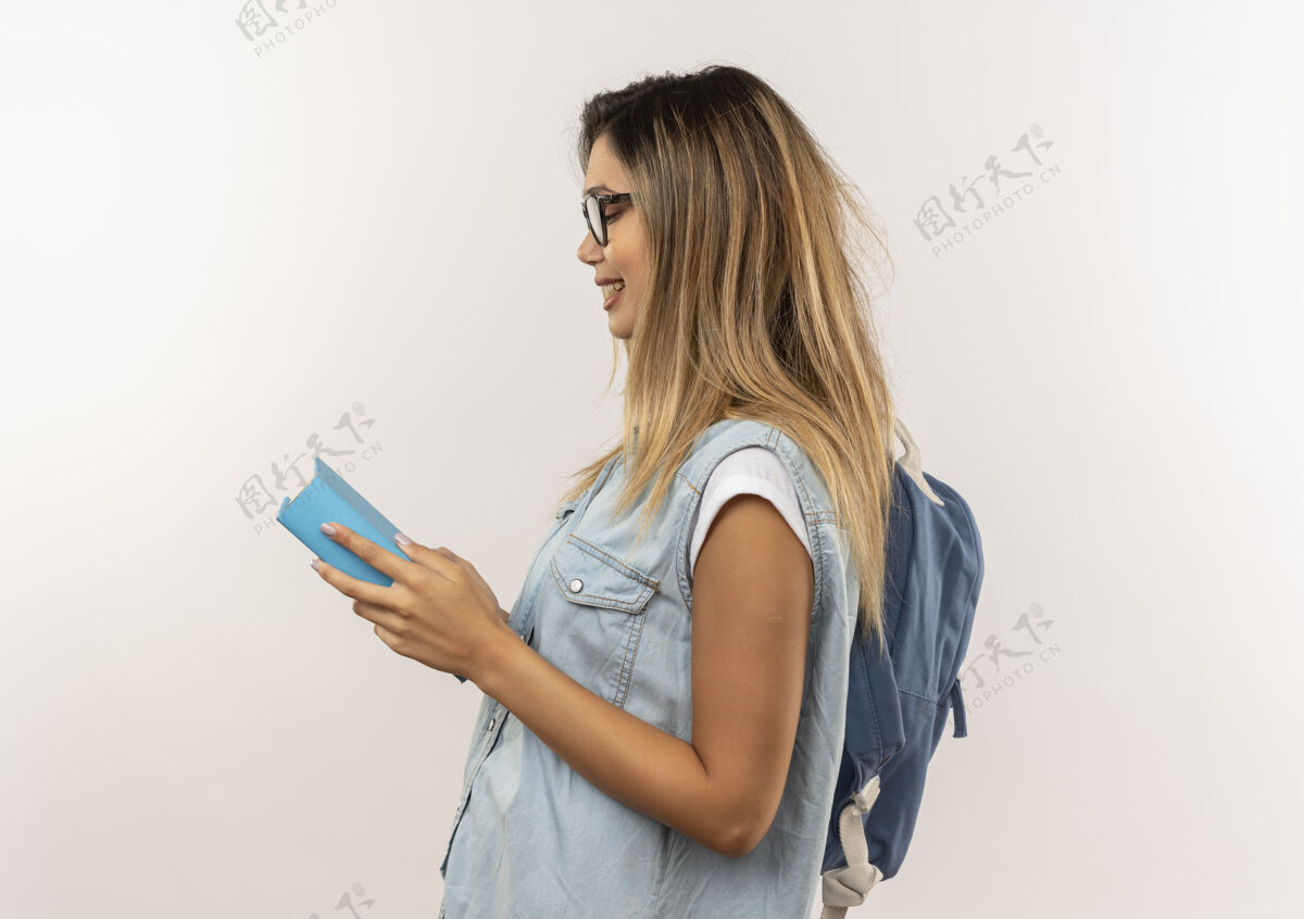 抱着面带微笑的年轻漂亮的女学生戴着眼镜 背着书包站在侧视图里 拿着书 看着隔离在白墙上的书眼镜脸站着