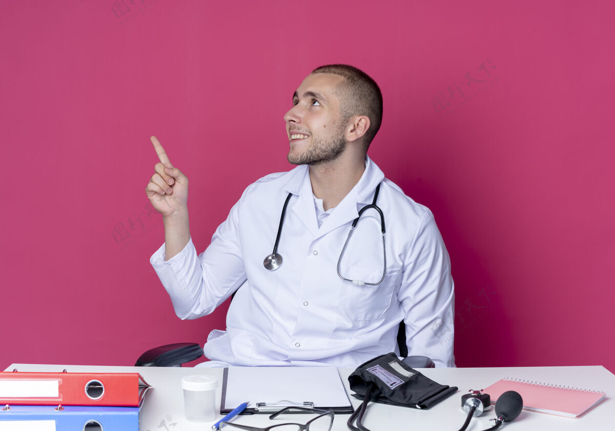 年轻人面带微笑的年轻男医生 穿着医用长袍 手持听诊器 坐在办公桌旁 手持工作工具 把头转向一边 孤立地指着粉红色的墙上感觉微笑脸