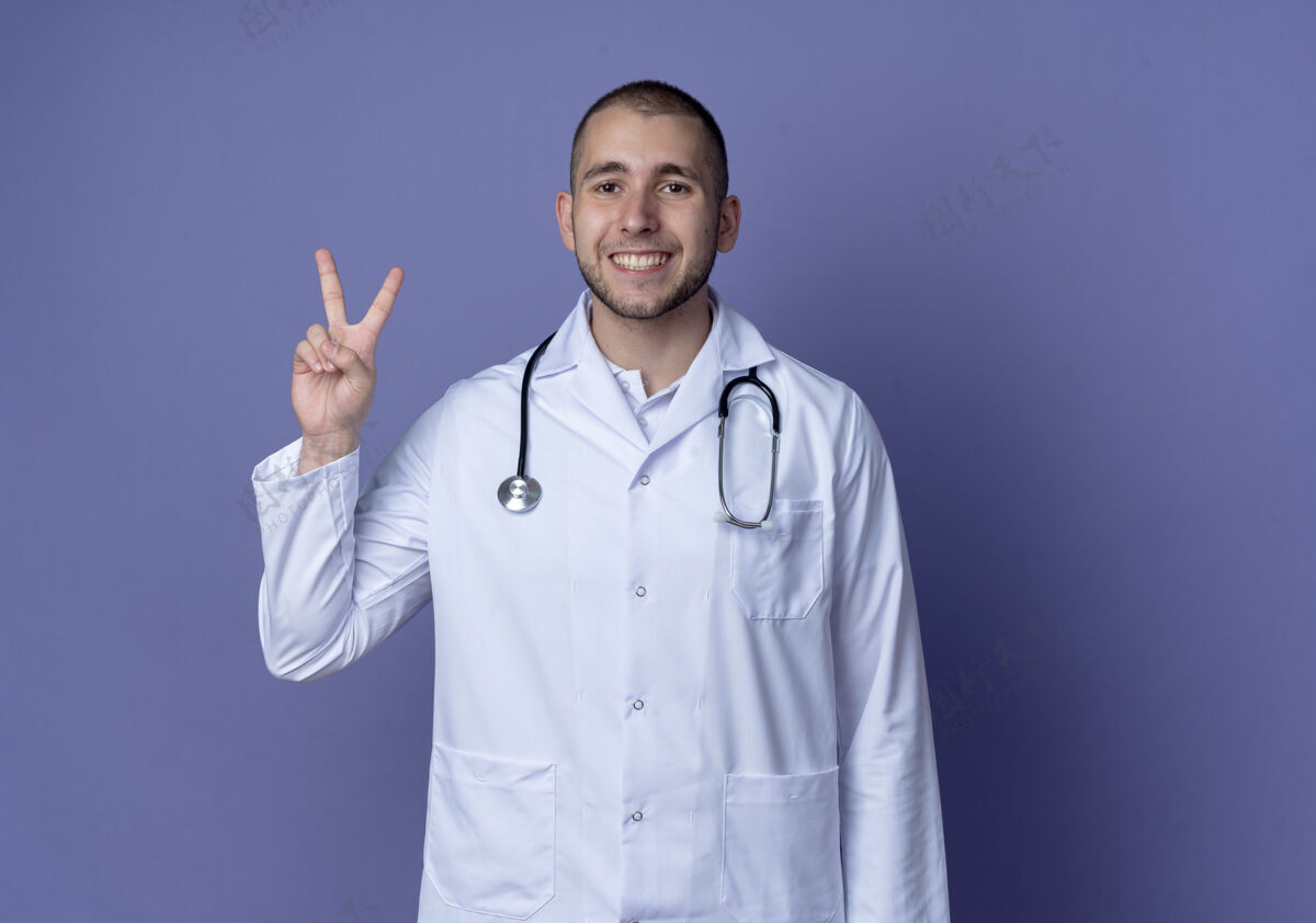 脸面带微笑的年轻男医生穿着医用长袍和听诊器做着和平的手势隔离在紫色的墙上医生衣服医学