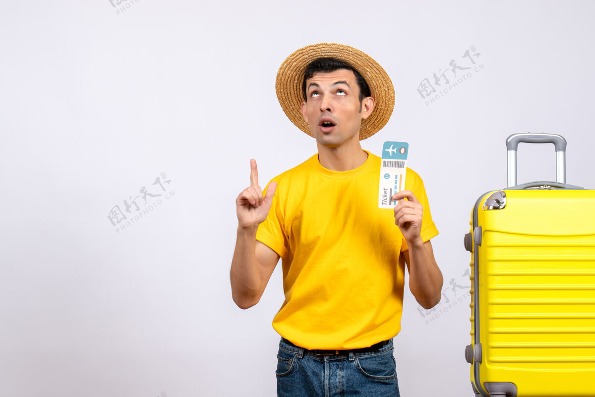男正面图穿着黄色t恤的年轻游客站在黄色手提箱旁 拿着票指着天花板手持T恤票