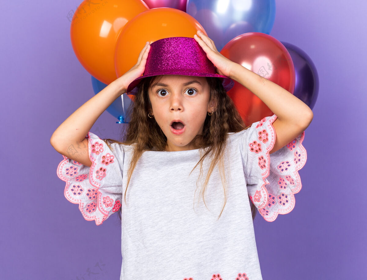 白种人震惊的小白种女孩 戴着紫色派对帽 手放在帽子上 站在氦气球前 在紫色的墙上 留着复制空间小孤立前面