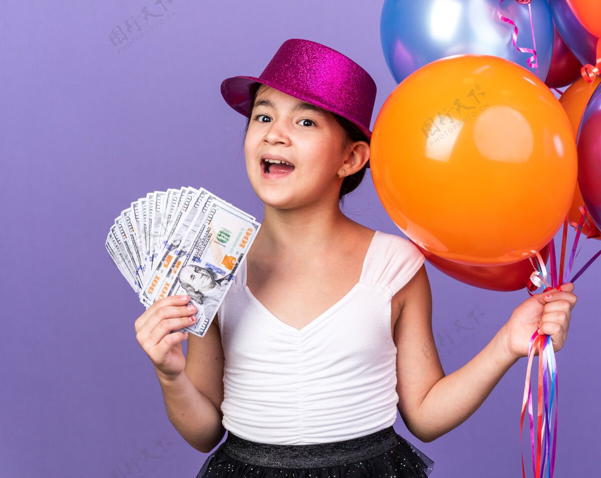 气球惊讶的年轻白种人女孩 戴着紫色派对帽 手里拿着氦气球 把钱孤立在紫色的墙上 留着复制空间帽子持有孤立