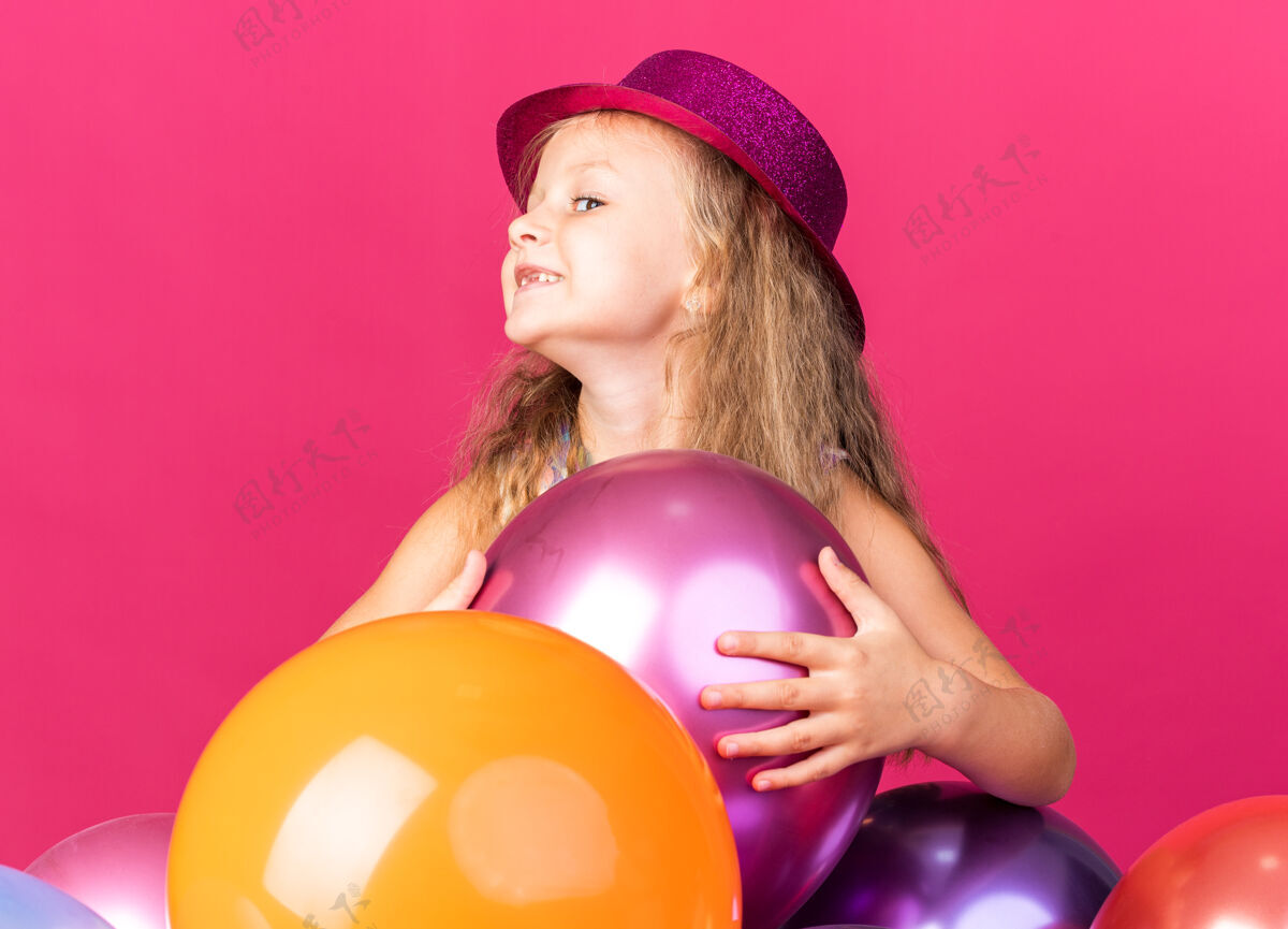 孤立微笑着的金发小女孩 戴着紫色派对帽 站着氦气球 看着粉色墙壁上孤立的一面 还有复制空间金发派对帽子