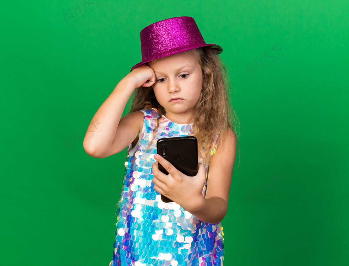 金发焦急的金发小女孩 戴着紫色的派对帽 看着隔离在绿色墙上的手机 还有复印空间聚会帽子生日