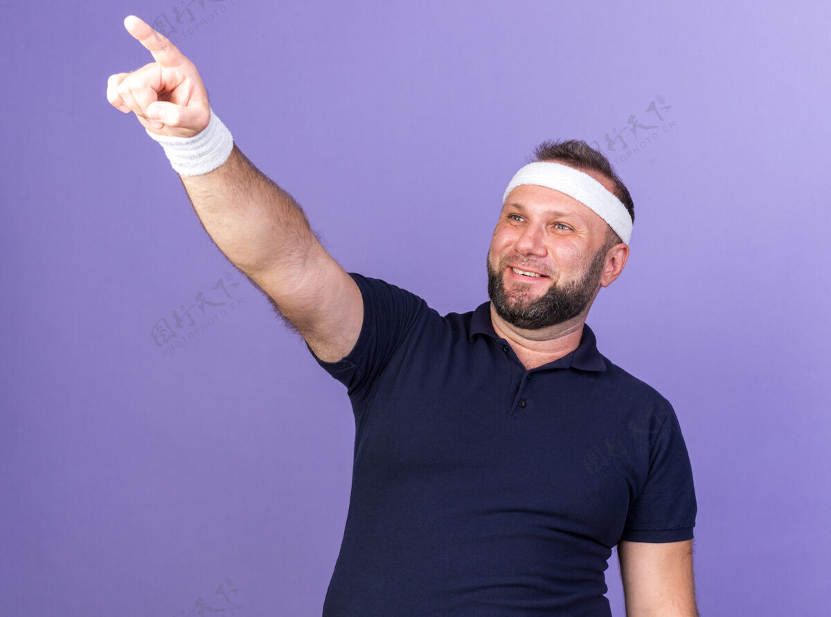 指点面带微笑的成年斯拉夫运动型男子戴着头带和腕带 看着并指着紫色墙上的复制空间孤立男人斯拉夫孤立