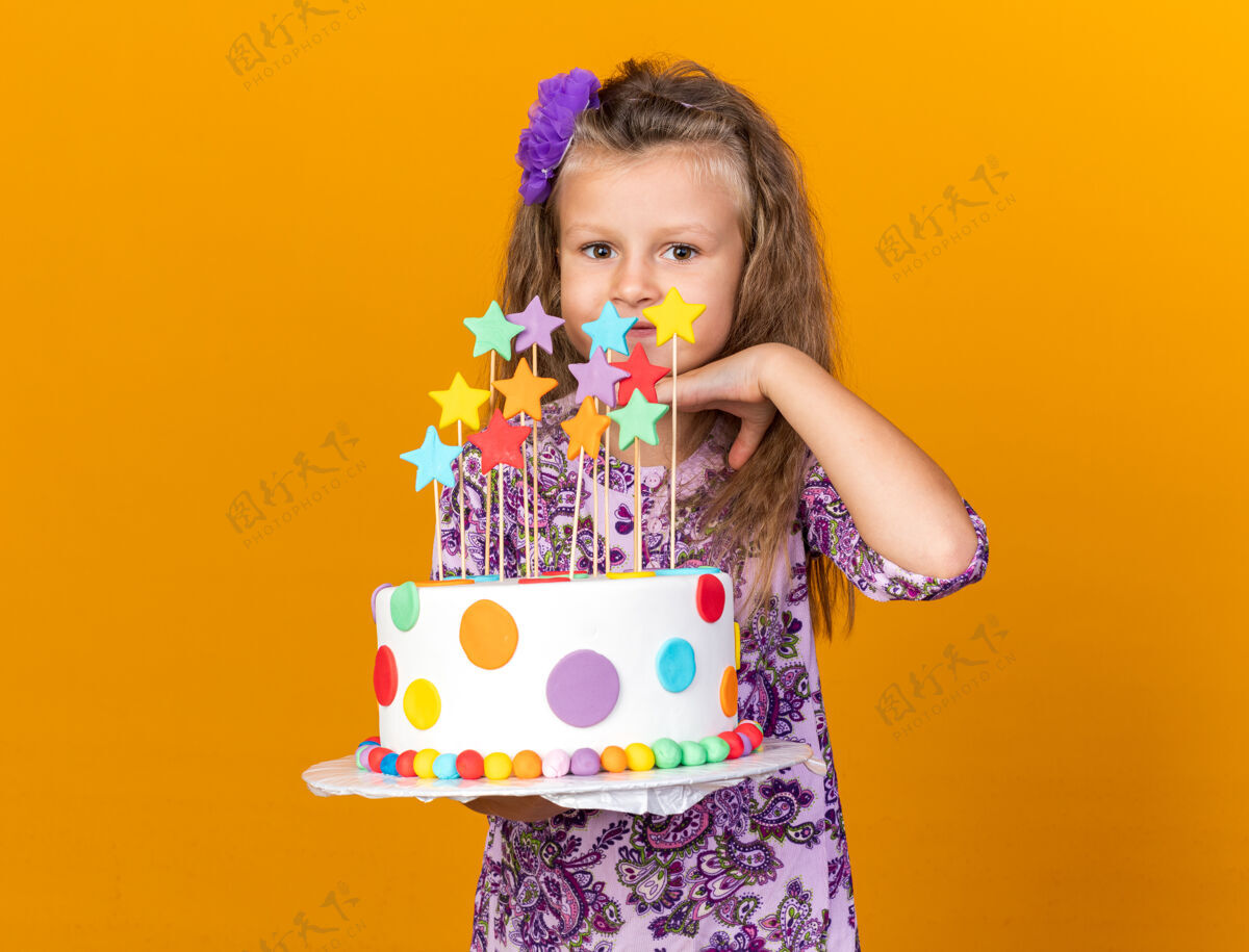 下巴高兴的金发小女孩拿着生日蛋糕 把手放在下巴上 隔离在橙色的墙上 留着复制空间抱着金发孤立