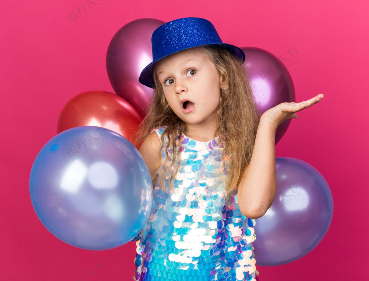 女孩惊讶的金发小女孩戴着蓝色的派对帽站在那里 手里拿着氦气球 手被隔离在粉色的墙上 还有复印空间打开帽子气球