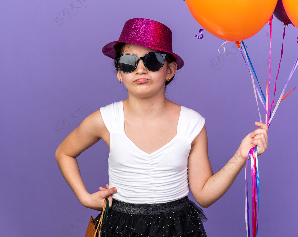 孤立戴着太阳眼镜 戴着紫色派对帽 拿着氦气球和购物袋的恼怒的白种年轻女孩被隔离在紫色的墙上 还有复印空间女孩包白种人