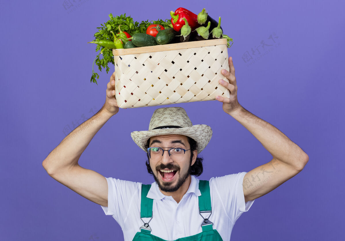 欢呼年轻的留着胡须的园丁 穿着连体衣 戴着帽子 头上顶着装满蔬菜的箱子 站在蓝色的墙上开心地微笑着蔬菜人人