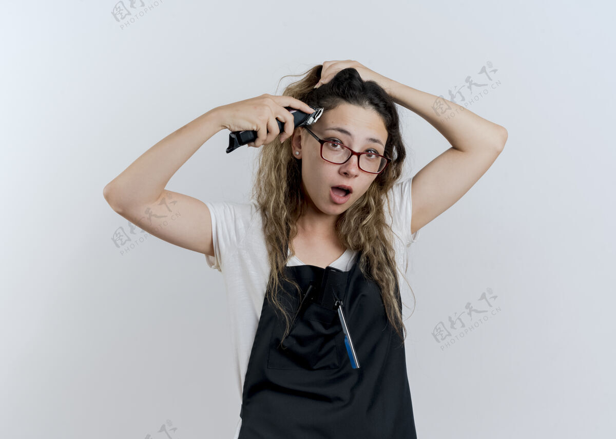 人年轻的专业美发师站在白色的墙上 围着围裙拿着修剪器试图剪头发 看上去很惊讶专业衣服头发