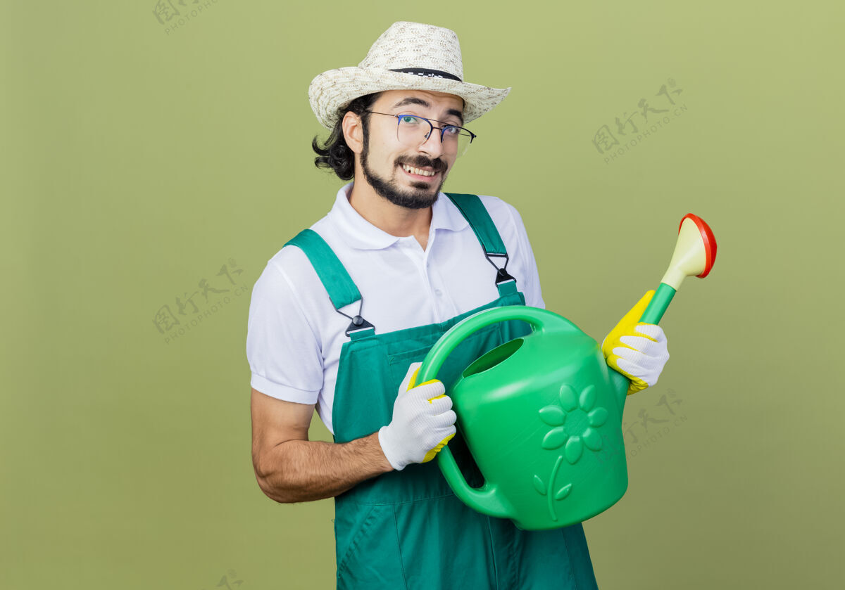 连身衣年轻的留着胡须的园丁 穿着连体衣 戴着帽子 手里拿着浇水罐 站在淡绿色的墙上 满脸笑容帽子胡须光