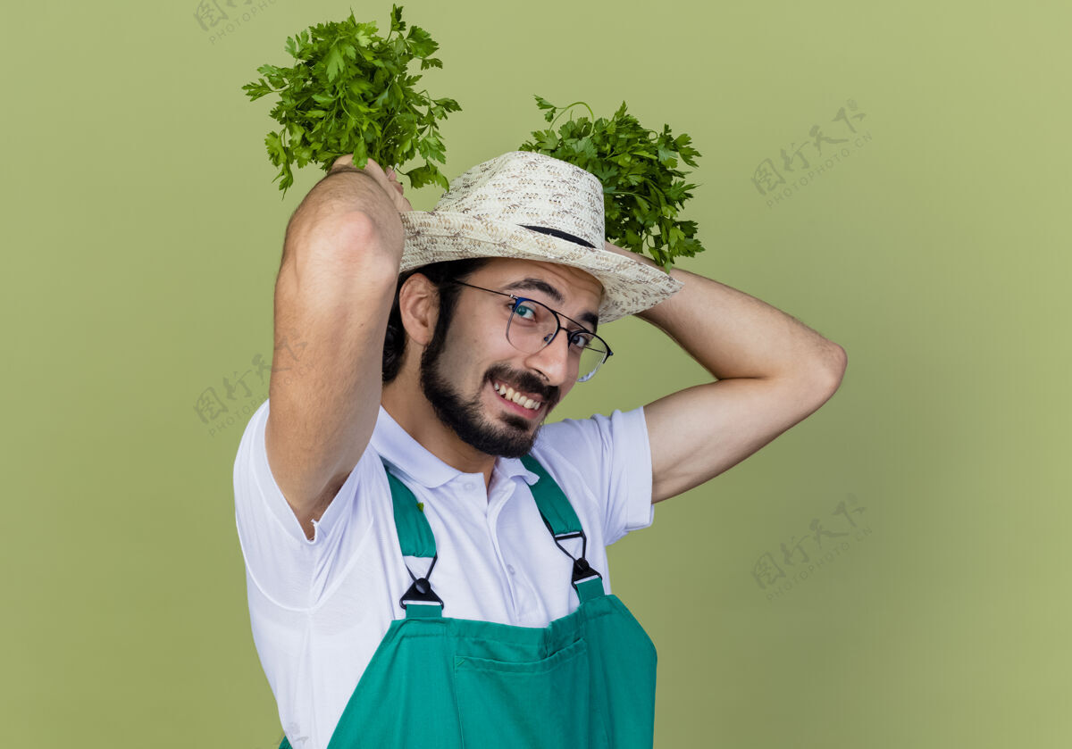 人有趣的年轻留着胡须的园丁 穿着连体衣 戴着帽子 手里拿着新鲜的草药 站在浅绿色的墙上 面带微笑地看着前面胡须感觉人