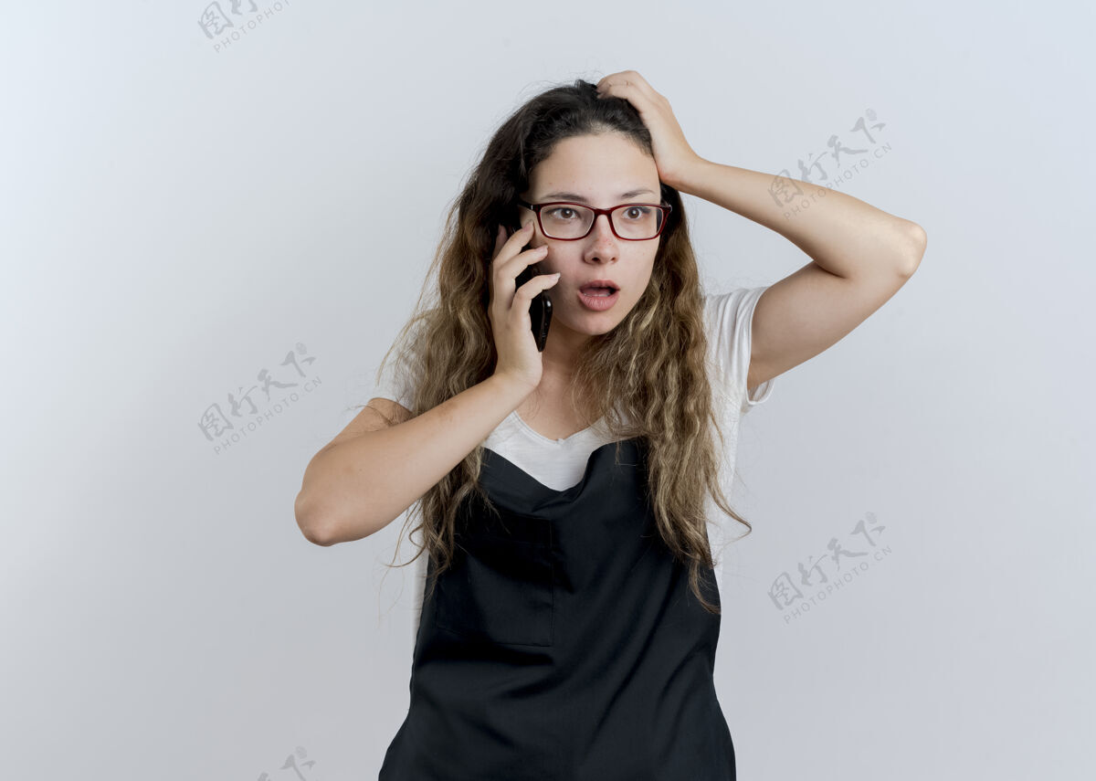 表情年轻的专业美发师围着围裙讲着手机站在白墙上困惑姿势女人衣服