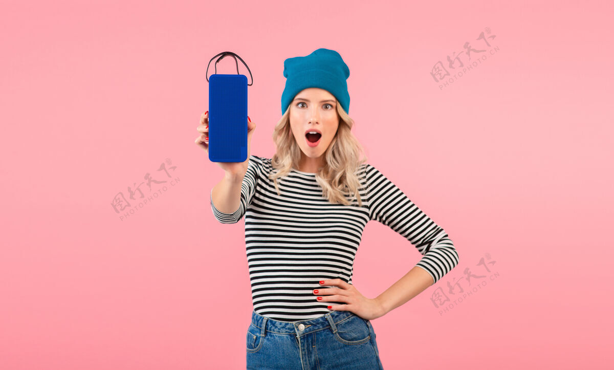 音乐年轻女子手持无线扬声器听音乐穿着条纹衬衫和蓝色帽子微笑着摆出粉红色的姿势女性享受穿着