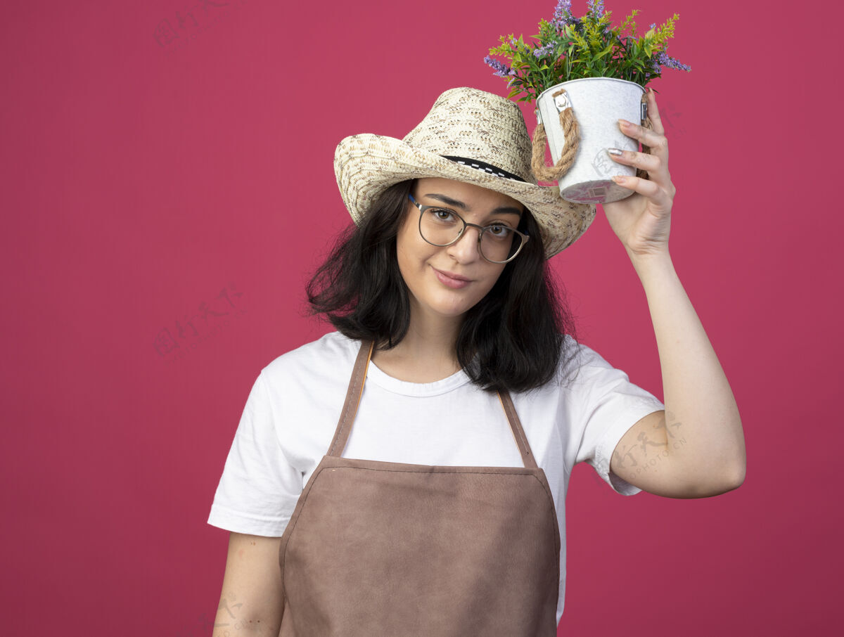 眼镜自信的年轻黑发女园丁戴着眼镜 身着制服 头戴园艺帽 将花盆举过头顶 隔离在粉红色的墙上穿着年轻人女性