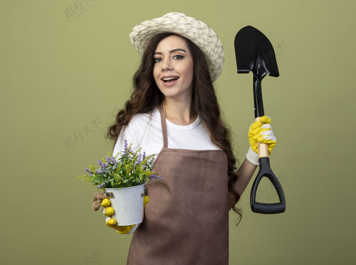 表情快乐的年轻女园丁穿着制服 戴着园艺帽和手套 手持铁锹和花盆 隔离在橄榄绿的墙上穿着花园脸