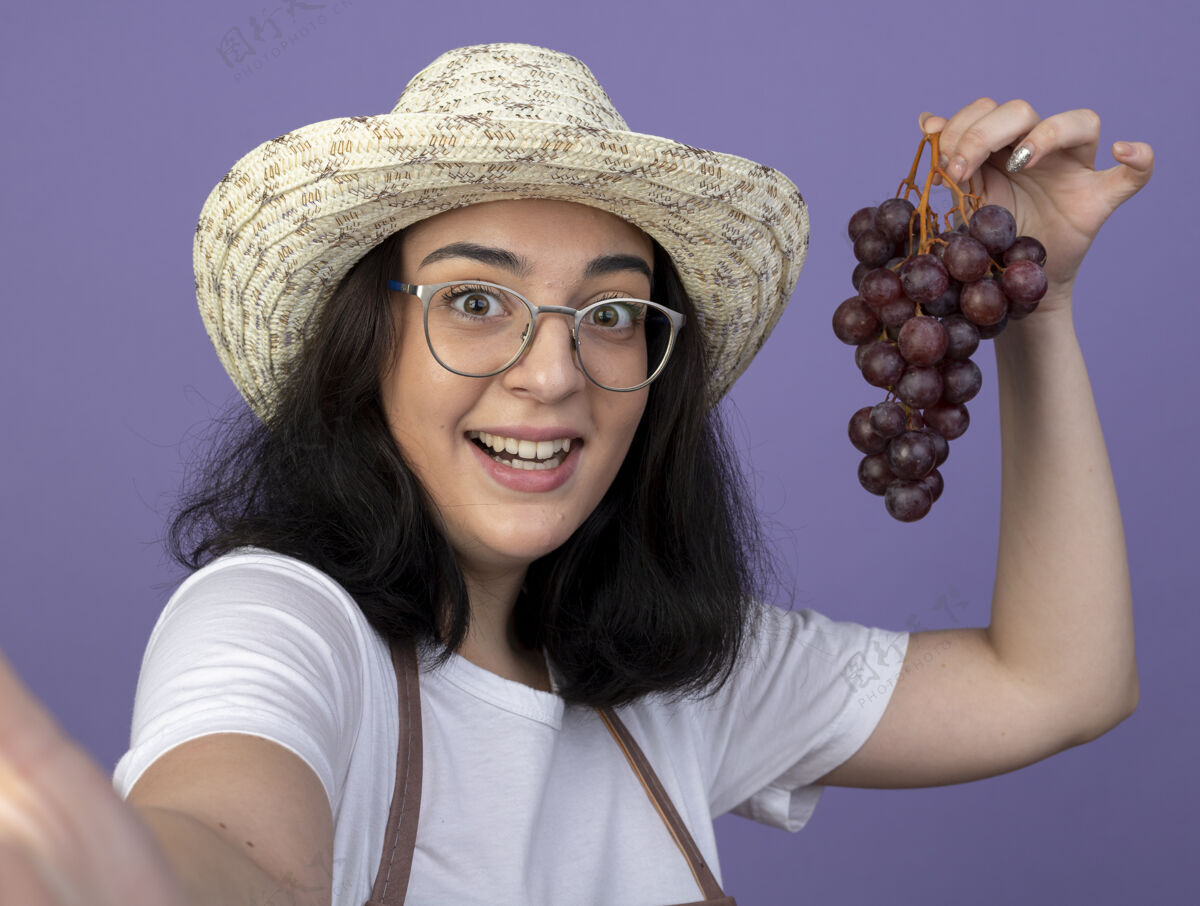 帽子惊讶的是 年轻的黑发女园丁戴着眼镜 穿着制服 戴着园艺帽 手里拿着葡萄 望着紫色墙壁上与世隔绝的前方人黑发制服