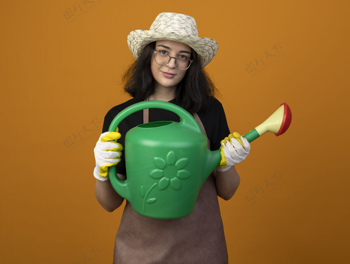 人高兴的是 年轻的黑发女园丁戴着眼镜 穿着制服 戴着园艺帽和手套 手里拿着隔离在橙色墙上的浇水罐人姿势拿着