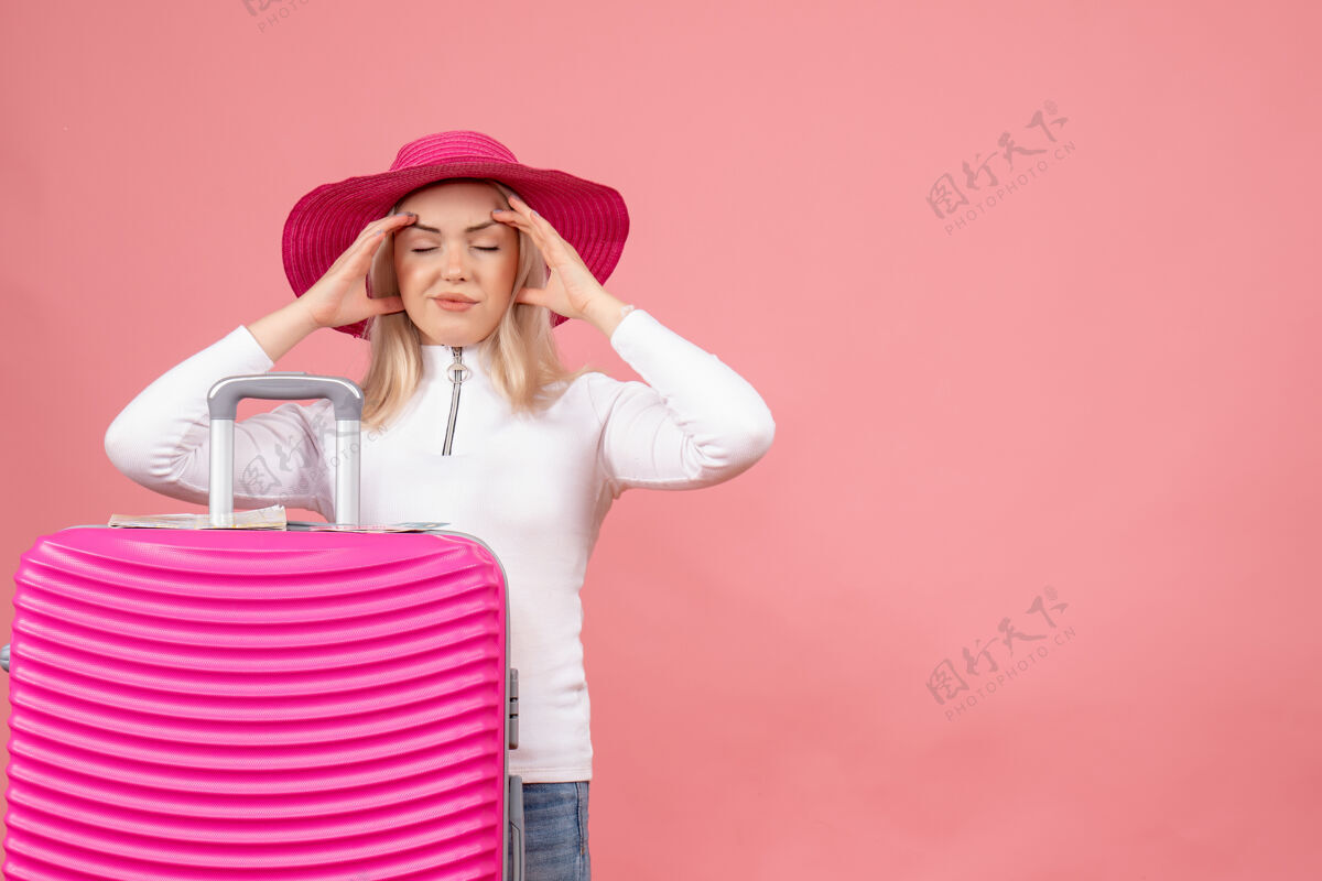 站立正面图：站在粉色手提箱后面的年轻女士抱着头前面衣服头