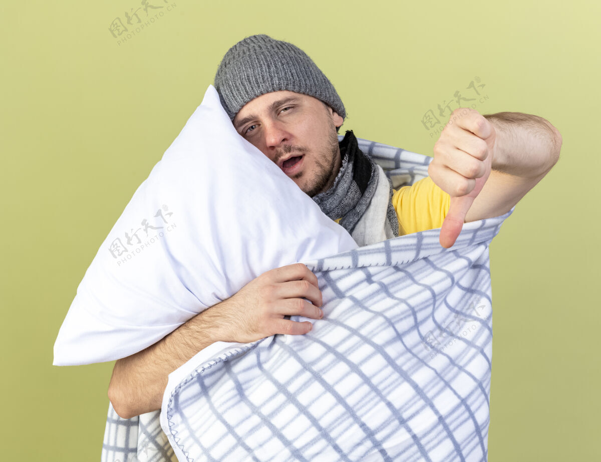 姿势苍白的年轻金发病男子戴着冬天的帽子裹着格子呢大拇指朝下 抱着橄榄绿墙上孤立的枕头包裹格子人