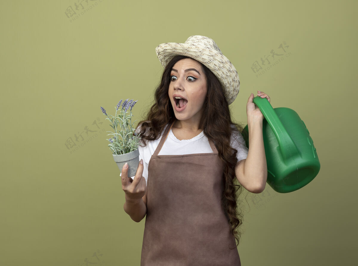 年轻穿着制服的年轻女园丁惊讶地戴着园艺帽 拿着喷壶 看着隔离在橄榄绿墙上的花盆里的花帽子举行穿着